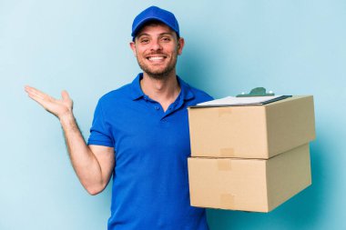 Beyaz tenli genç teslimatçı mavi arka planda izole edilmiş bir şekilde bir avuç içinde fotokopi alanı ve belinde bir el daha gösteriyor..