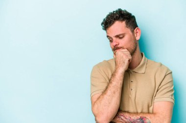 Mavi arka planda izole edilmiş genç beyaz adam şüpheli ve şüpheci bir ifadeyle yana bakıyor..