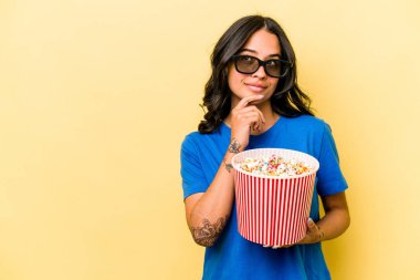 Sarı arka planda izole edilmiş patlamış mısır tutan genç İspanyol kadın şüpheli ve şüpheci bir ifadeyle yana bakıyor..