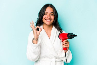 Mavi arka planda izole edilmiş kurutma makinesini tutan genç İspanyol kadın neşeli ve kendinden emin..