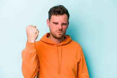 Mavi arka planda izole edilmiş genç beyaz adam yumruğunu kameraya gösteriyor, agresif yüz ifadesi var..