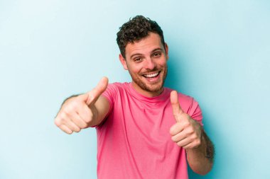 Kafkasyalı genç adam mavi arka planda izole edilmiş. Baş parmağını kaldırıyor, gülümsüyor ve kendinden emin..