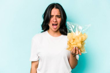 Mavi arka planda izole edilmiş bir paket cipsi tutan genç İspanyol kadın çok sinirli ve agresif bağırıyor..