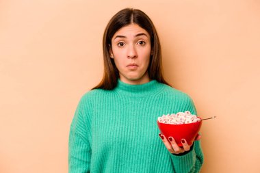 Genç İspanyol kadın bej arka planda izole mısır gevreği yiyor omuzlarını silkiyor ve gözleri açık..