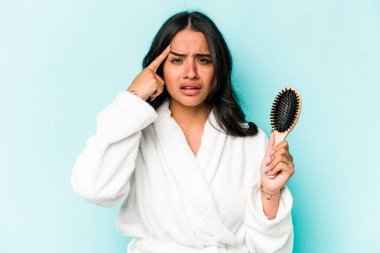 Mavi arka planda saç fırçası tutan genç İspanyol kadın işaret parmağıyla yaptığı hayal kırıklığı jestini gösteriyor..