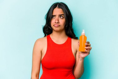 Mavi arka planda güneş kremi tutan genç İspanyol kadın şaşkın ve kararsız hissediyor..