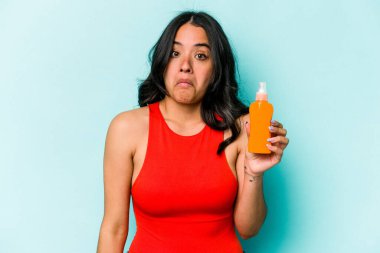 Mavi arka planda izole edilmiş güneş kremi tutan genç İspanyol kadın omuzlarını silker ve gözleri açık kalır..