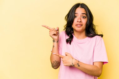 Sarı arka planda izole edilmiş genç İspanyol kadın işaret parmağıyla fotokopi alanını işaret ederek şok oldu..