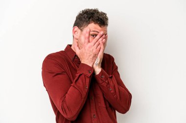 Beyaz arka planda izole edilmiş genç beyaz bir adam parmaklarının arasından korkmuş ve gergin bir şekilde göz kırpıyor..