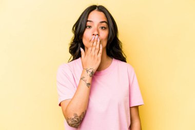 Sarı arka planda izole edilmiş genç İspanyol kadın şok olmuş, ağzı ellerle kaplanmış, yeni bir şey keşfetmeye hevesli..