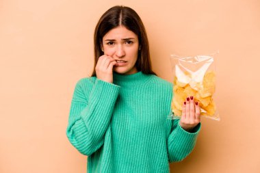 Latin kökenli genç bir kadın elinde bir çanta dolusu cips tutuyor. Bej arka planda izole edilmiş tırnak yiyor, gergin ve endişeli..