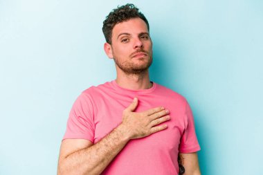 Kafkasyalı genç adam mavi arka planda izole edilmiş yemin ediyor, elini göğsüne koyuyor..