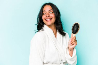 Mavi arka planda saç fırçası tutan genç İspanyol kadın gülümsüyor, neşeli ve keyifli görünüyor..