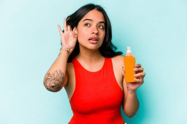 Mavi arka planda güneş kremi tutan genç İspanyol kadın bir dedikoduyu dinlemeye çalışıyor..