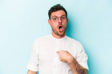 Mavi arka planda izole edilmiş genç beyaz adam parmağını göstererek şaşırdı, geniş bir gülümsemeyle..