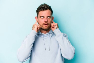 Genç beyaz adam mavi arka planda izole edilmiş parmaklarıyla kulaklarını kapatıyor, gürültülü bir ortam tarafından stresli ve umutsuz..