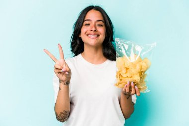 Mavi arka planda iki numarayı parmaklarıyla gösteren bir paket cipsi tutan genç İspanyol kadın..