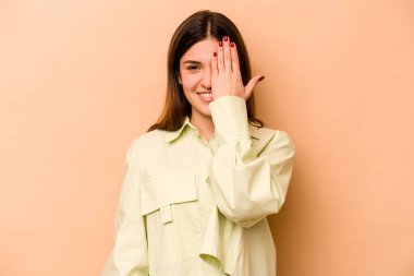 Genç İspanyol kadın bej arka planda izole edilmiş. Yüzünün yarısını avuç içiyle kaplayarak eğleniyor..