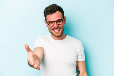 Mavi arka planda izole edilmiş genç beyaz adam selamlama jesti olarak kameraya elini uzatıyor..