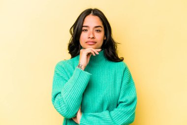 Sarı arka planda izole edilmiş genç İspanyol kadın şüpheli, belirsiz, seni muayene ediyor..