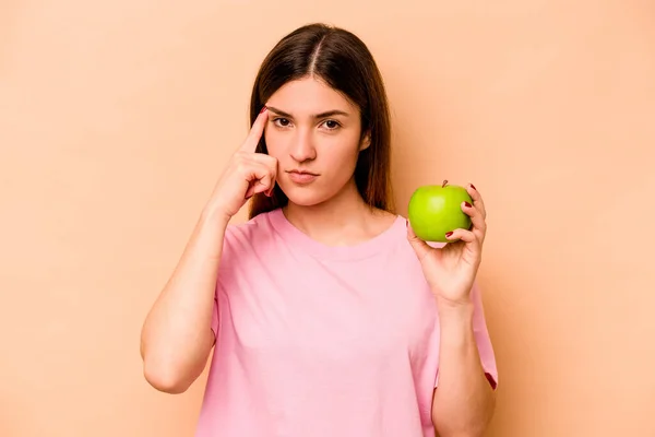 Ung Latinamerikansk Kvinna Håller Ett Äpple Isolerat Beige Bakgrund Pekar — Stockfoto