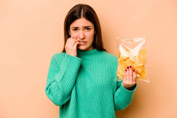 Latin kökenli genç bir kadın elinde bir çanta dolusu cips tutuyor. Bej arka planda izole edilmiş tırnak yiyor, gergin ve endişeli..