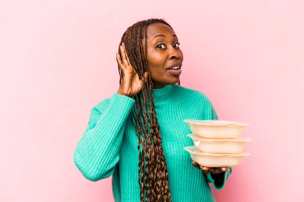 Young African American woman holding tupperware isolated on pink background trying to listening a gossip.