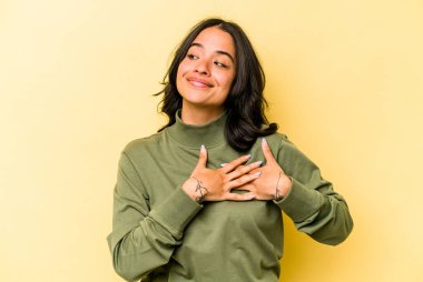 Sarı arka planda izole edilmiş genç İspanyol kadın, dost canlısı bir ifadeye sahip, avuçlarını göğsüne bastırıyor. Aşk konsepti.