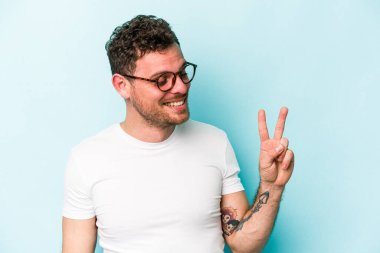 Kafkasyalı genç adam mavi arka planda izole edilmiş neşeli ve kaygısız parmaklarıyla barış sembolü gösteriyor..
