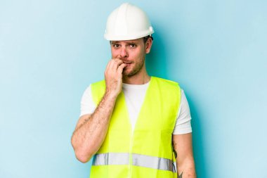 Kafkasyalı genç işçi, mavi arka planda izole edilmiş tırnak yiyor, gergin ve endişeli..