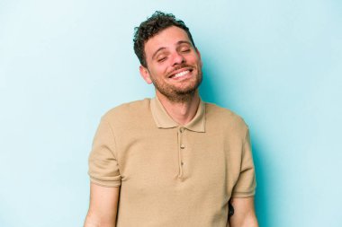 Mavi arka planda izole edilmiş genç beyaz adam gülüyor ve gözlerini kapıyor, rahatlamış ve mutlu hissediyor..