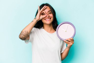 Mavi arka planda izole edilmiş bir saat tutan genç İspanyol kadın heyecanlı..