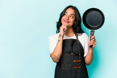 Genç İspanyol tencereli kadın elinde kızartma tavasıyla mavi arka planda izole edilmiş şüpheli ve şüpheci bir ifadeyle yana bakıyor..