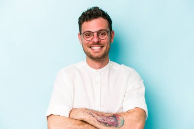 Mavi arka planda kendini güvende hisseden, kararlılıkla kollarını kavuşturan genç beyaz bir adam..