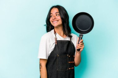 Mavi arka planda izole edilmiş tavayı tutan İspanyol tencereli genç kadın gülümsüyor, neşeli ve keyifli görünüyor..