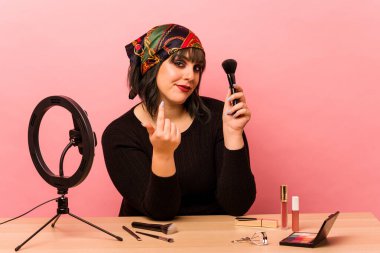 Makyaj sanatçısı genç bir kadın pembe arka planda izole edilmiş bir makyaj dersi veriyor. Sanki sizi davet ediyormuş gibi parmağıyla işaret ediyor..