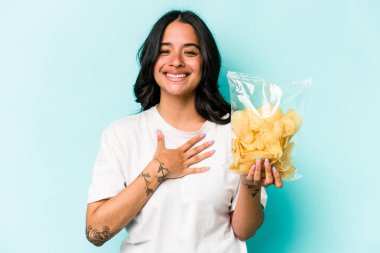 Mavi arka planda izole edilmiş bir paket cipsi tutan genç İspanyol kadın kahkahalarla gülüyor..