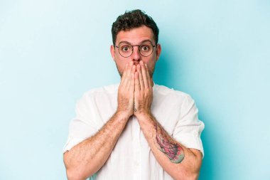 Mavi arka planda izole edilmiş genç beyaz adam elleriyle ağzını kapatırken şok oldu..
