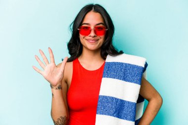 Mavi arka planda izole edilmiş plaj havlusu tutan genç İspanyol kadın gülümseyerek beş numarayı parmaklarıyla gösteriyor..