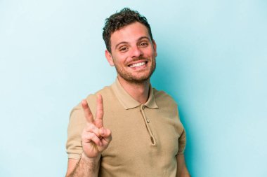 Mavi arka planda izole edilmiş genç beyaz adam zafer işareti gösteriyor ve gülümsüyor..