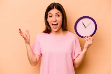 Latin kökenli genç bir kadın bej arka planda izole edilmiş bir saat tutuyor. Hoş bir sürpriz alıyor, heyecanlı ve el kaldırıyor..