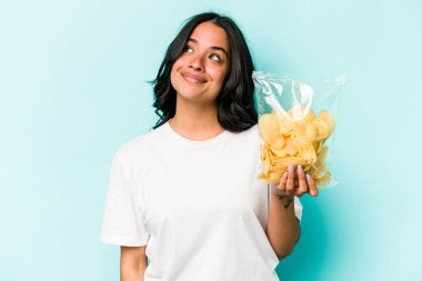 Genç İspanyol kadın elinde bir çanta dolusu cipsle mavi arka planda izole bir şekilde amaçlarına ve amaçlarına ulaşmayı hayal ediyor.