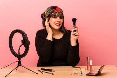 Makyaj eğitmeni genç bir kadın pembe arka planda izole edilmiş elleriyle kulaklarını kapatıyor..