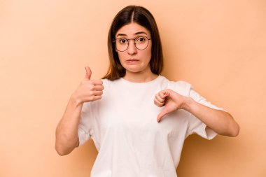 Latin kökenli genç bir kadın bej arka planda izole edilmiş. Baş parmağını kaldırmış, baş parmağını aşağı gösteriyor. Zor seçim konsepti.