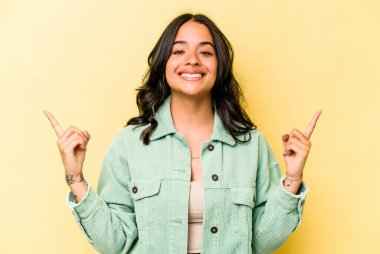 Sarı arka planda izole edilmiş genç İspanyol kadın iki parmağı da boş bir alan gösteriyor..