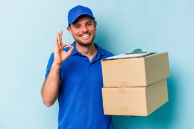 Beyaz tenli genç teslimatçı mavi arka planda izole edilmiş neşeli ve kendinden emin bir şekilde iyi niyet gösteriyor..