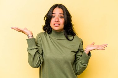 Sarı arka planda izole edilmiş genç İspanyol kadın. Kafası karışmış ve omuz silkiyor. Kopya alanını tutmak için..