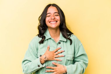 Sarı arka planda izole edilmiş genç İspanyol kadın mutlu bir şekilde gülüyor ve ellerini karnının üstünde tutarak eğleniyor..