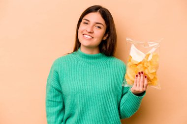 Genç İspanyol kadın elinde bir paket cipsle bej arka planda izole edilmiş mutlu, güler yüzlü ve neşeli.