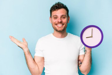 Young caucasian man holding a clock isolated on blue background showing a copy space on a palm and holding another hand on waist.